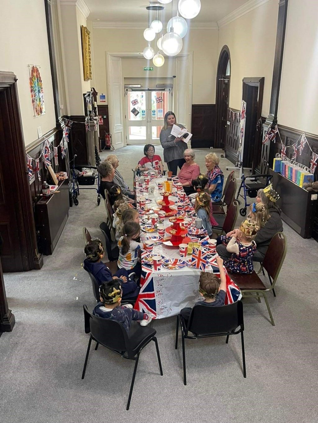 A group of children and adults enjoying a lunch to celebrate the King's coronation. The table is decorated with union jack flags and most people are wearing paper crowns.