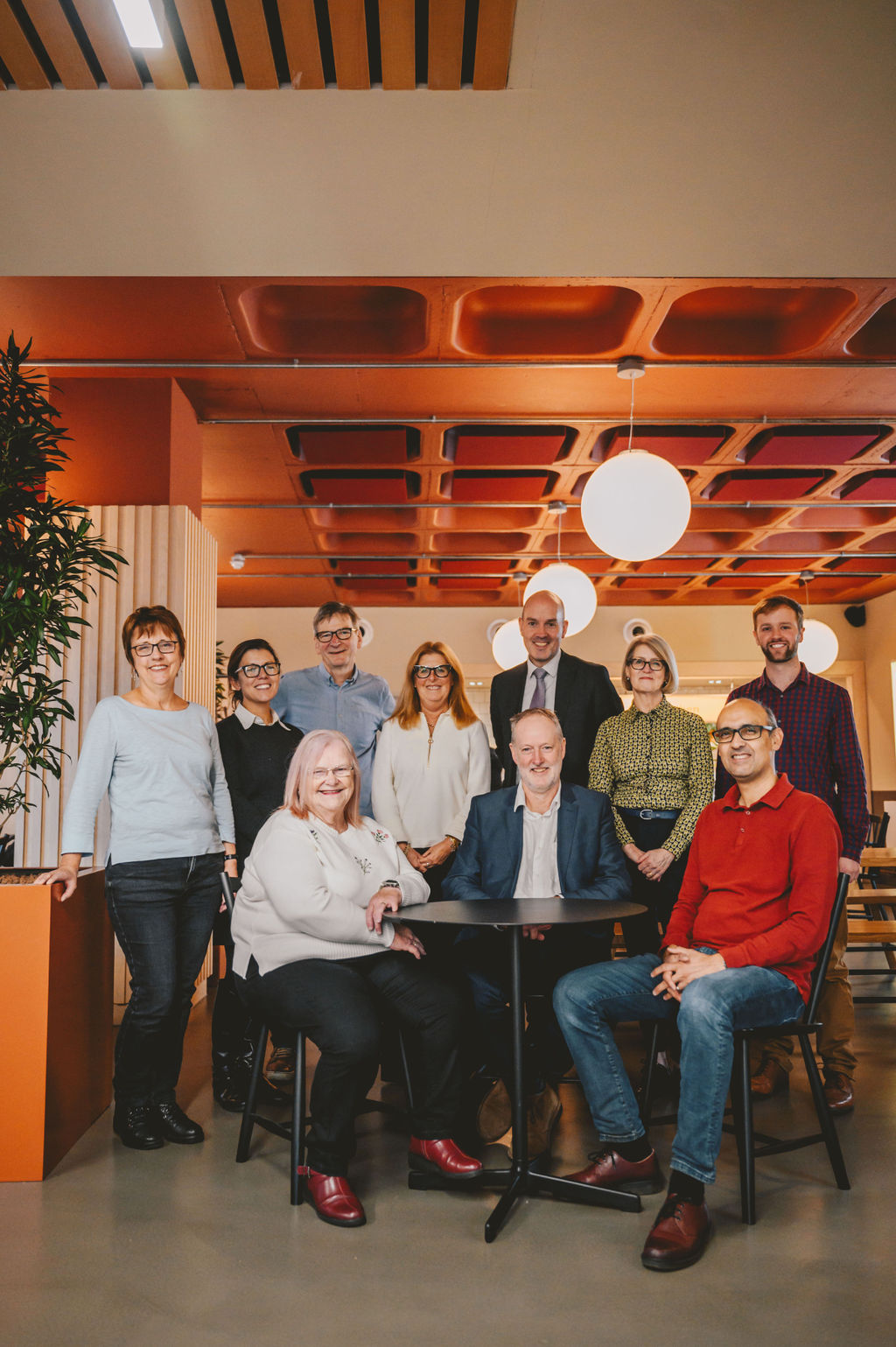 A photo of some of our board members. Some are sat around a table, and others are stood next to and behind it. They are all looking at the camera and smiling.