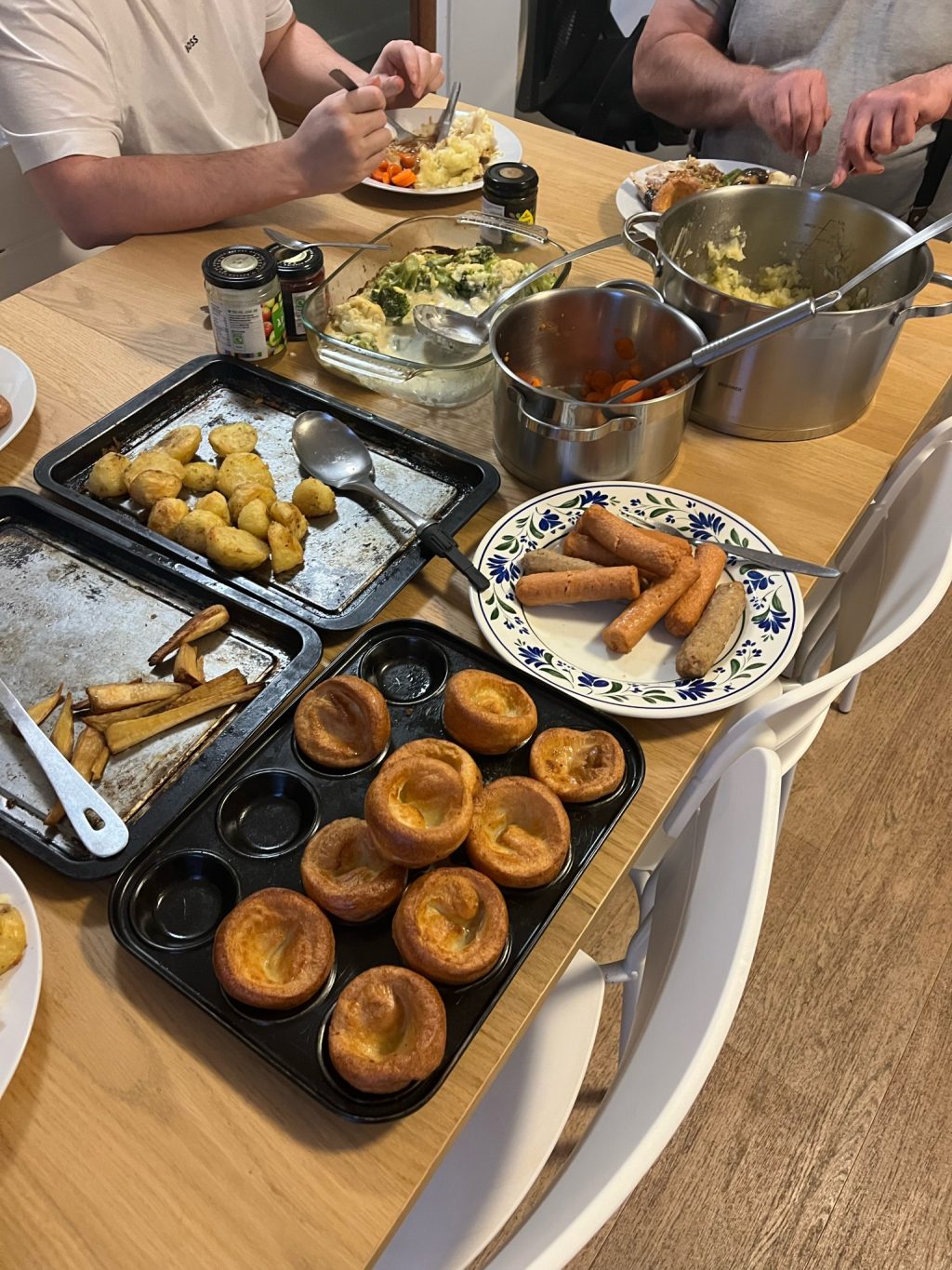 A roast dinner at the dinner table, including a tray of yorkshire puddings, some potatoes, and some plates of vegetables. You can see the plates and hands of people enjoying their food.