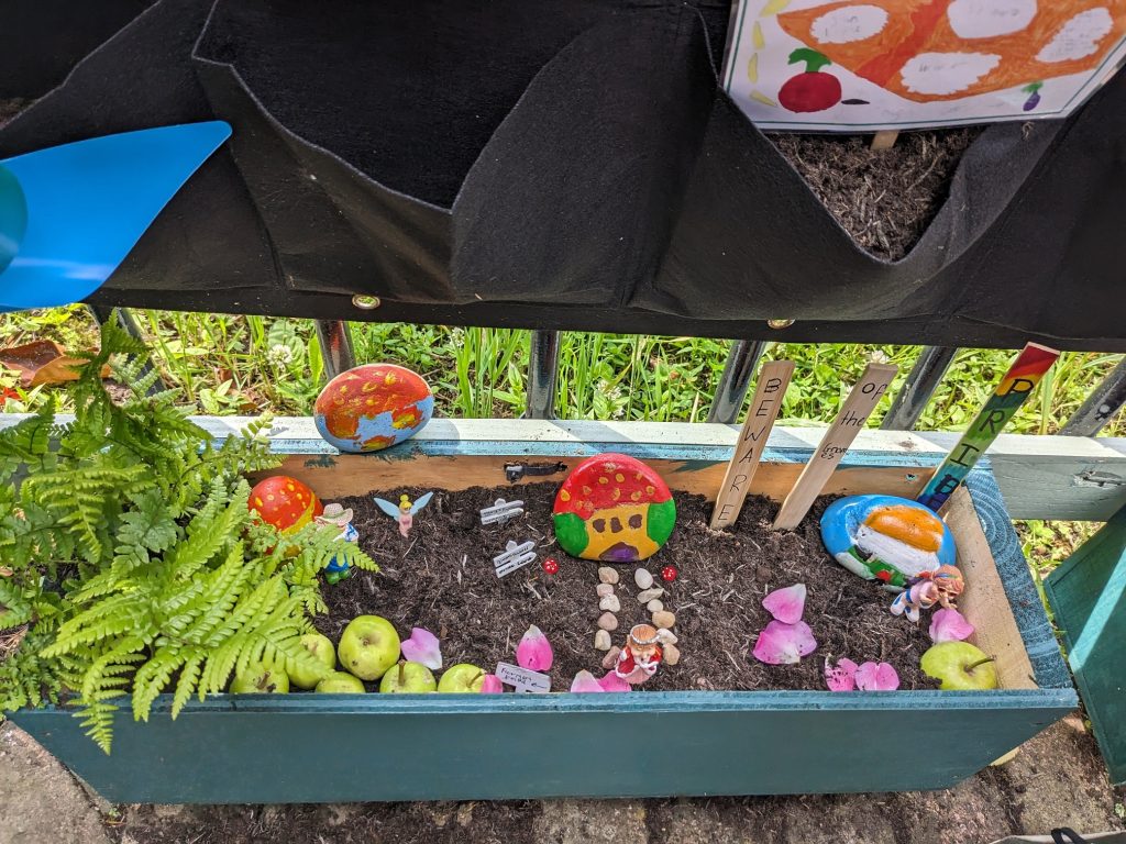 A garden planter filled with brightly coloured signs, pebbles, petals, plants and apples.