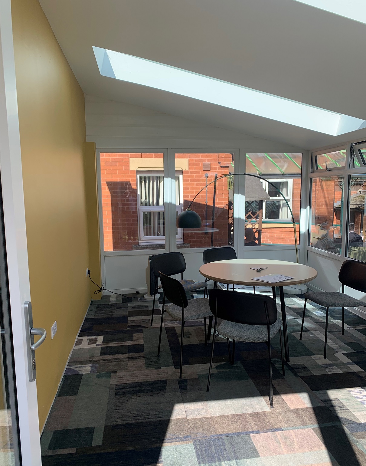 A view of the summer house - a bright room with a yellow painted wall, with a table, chairs, lamp and big windows.