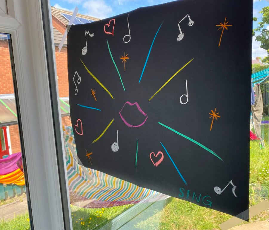 Artwork on the window of the Lister summer house - colourful shapes and lines on black paper, with a pink mouth and the words 'sing'.