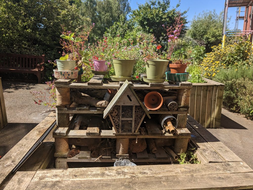 A picture of a bug hotel set within a handmade wooden shelving system among gardening pots in the garden at The Meadows