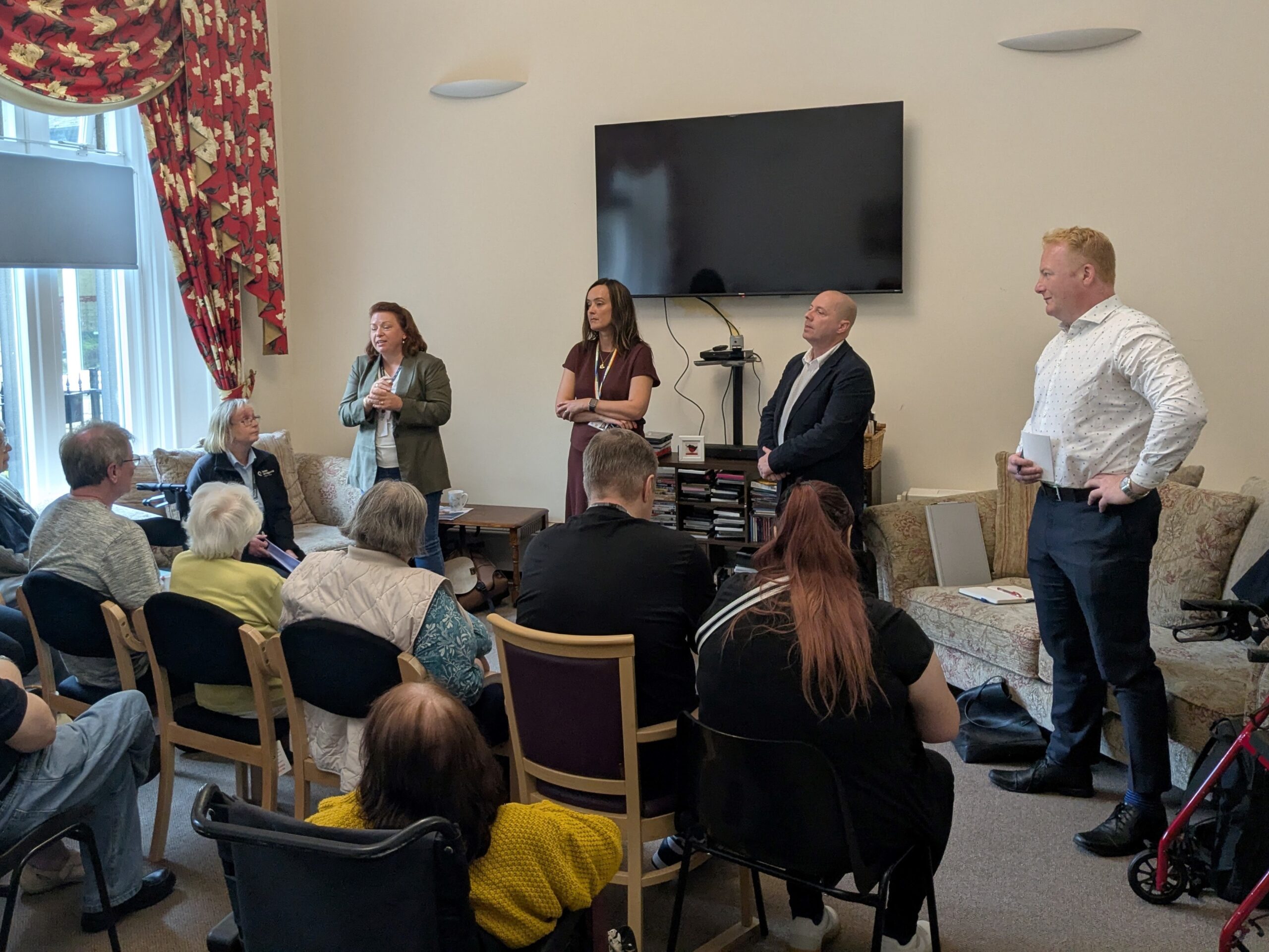 A photo of one of our customer consultation events. Four people are stood talking to an seated audience.