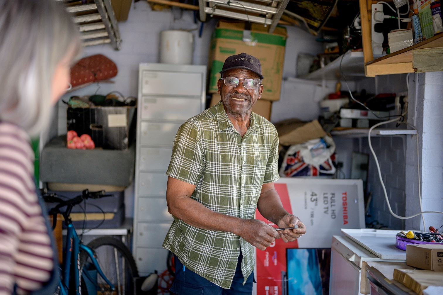 An older man standing in his shed and smiling. He's wearing a black hat and a short sleeved green shirt.